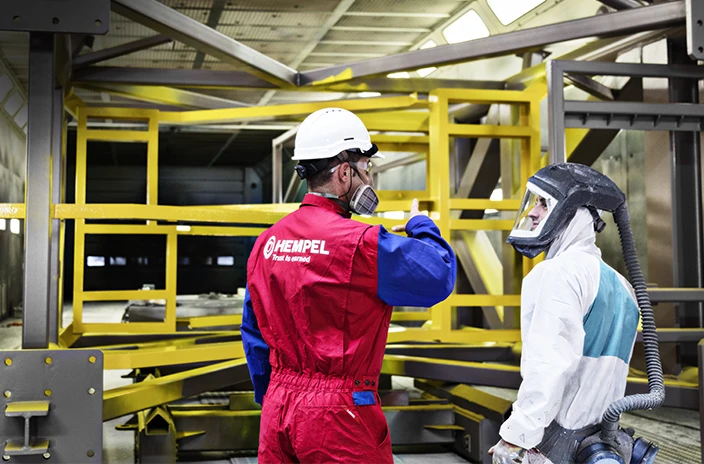 A coating advisor wearing a red and blue Hempel uniform speaks to a worker inside a large industrial structure.