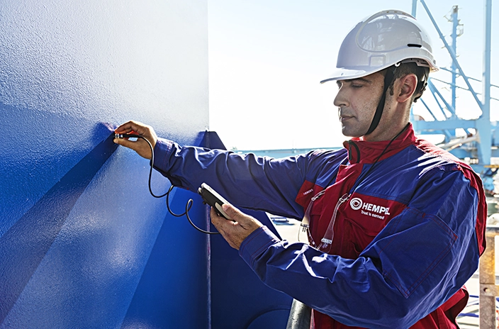 A coating advisor from Hempel measuring coating thickness on a blue painted surface.