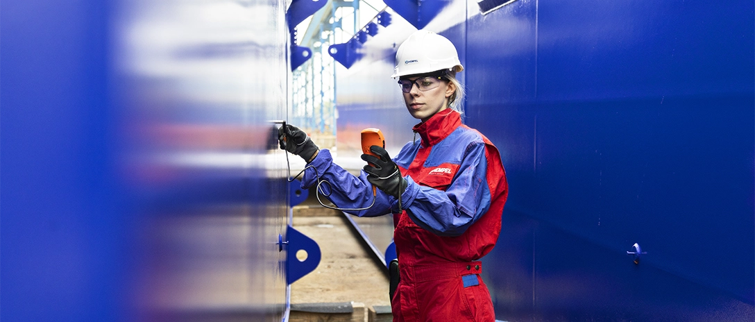 A Hempel female coating advisor measures a blue surface with a handheld device, standing inside a large industrial structure.