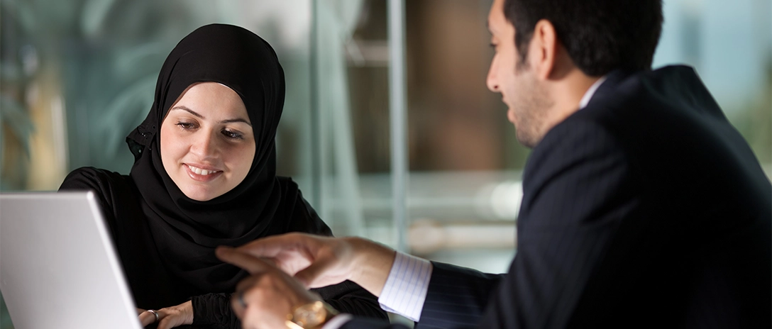 Two people in office engaged in a discussion while reviewing something on a laptop.