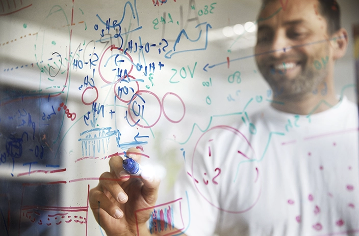 A man in a laboratory writes a strategy map with equations and graphs on a glass board, highlighting research and development work.