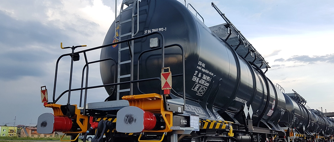 A close-up view of a black tanker railcar, equipped with ladders and safety markings, lined up with other tankers under a cloudy sky.