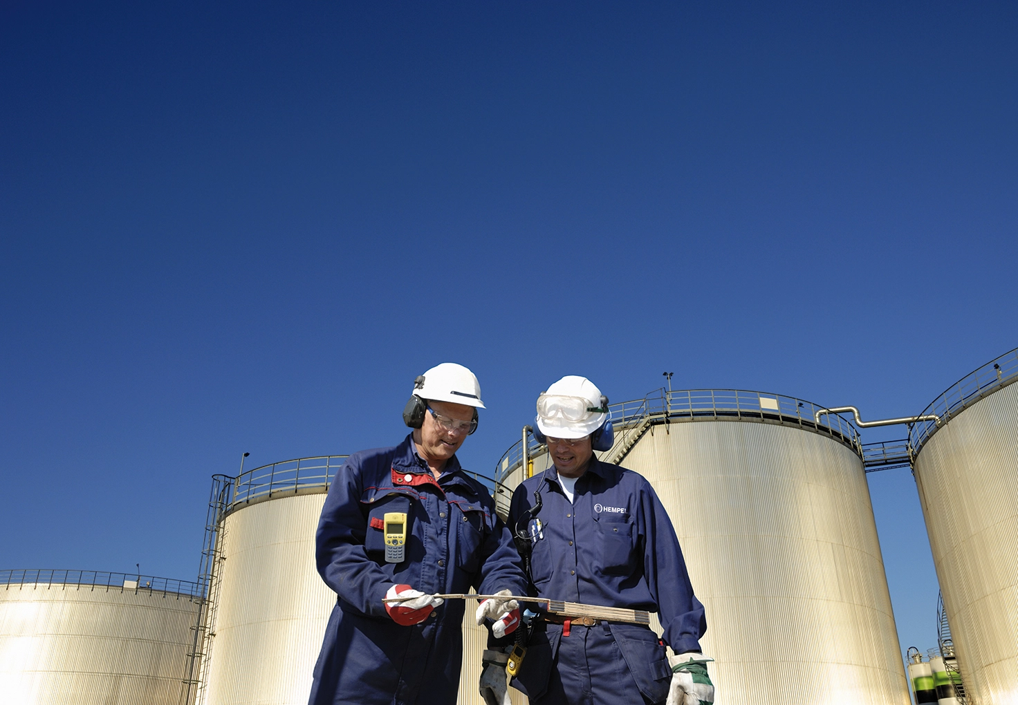 Two workers in blue uniforms review a wooden scale for measuring, with large storage tanks in the background under a clear sky.