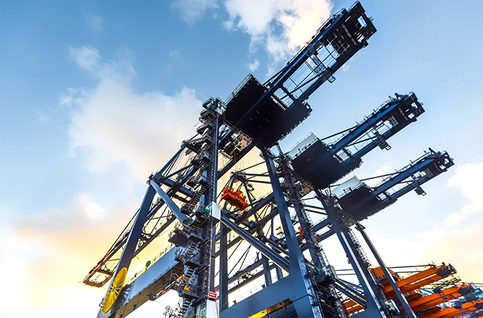 A close-up view of large container cranes at a port, silhouetted against a bright sky with clouds.