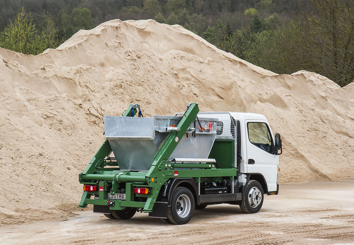 White Jotha Fahrzeugbau truck with a green hydraulic lift carrying a metal container, parked in a sandy industrial area.